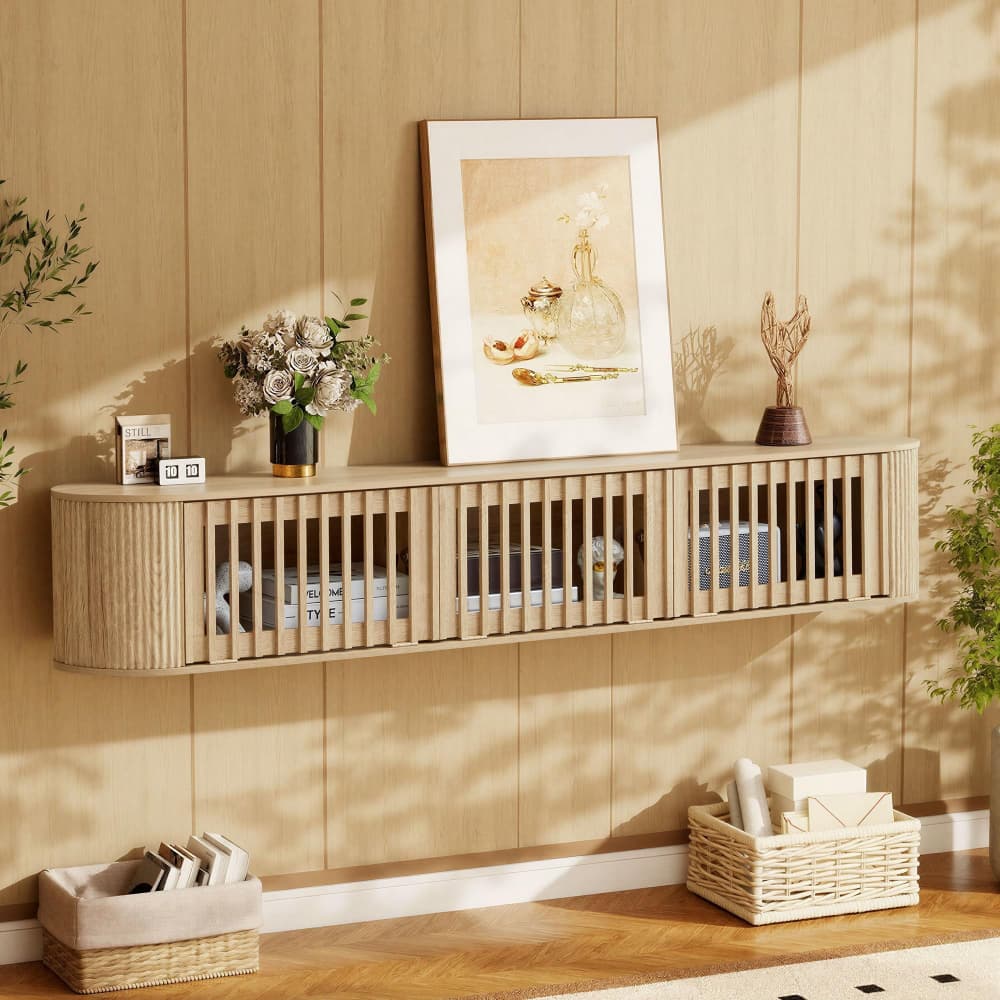 A light natural wood floating wall cabinet with vertical slat detailing, styled in a warm, sunlit room against a wood-paneled wall with a framed print, flowers, and baskets.