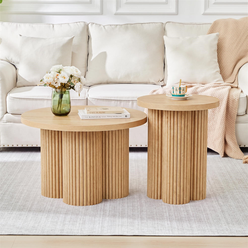 Two round wooden coffee tables in a bright living room. One table has a fluted pedestal base and holds a green glass vase with white flowers and a stack of books.