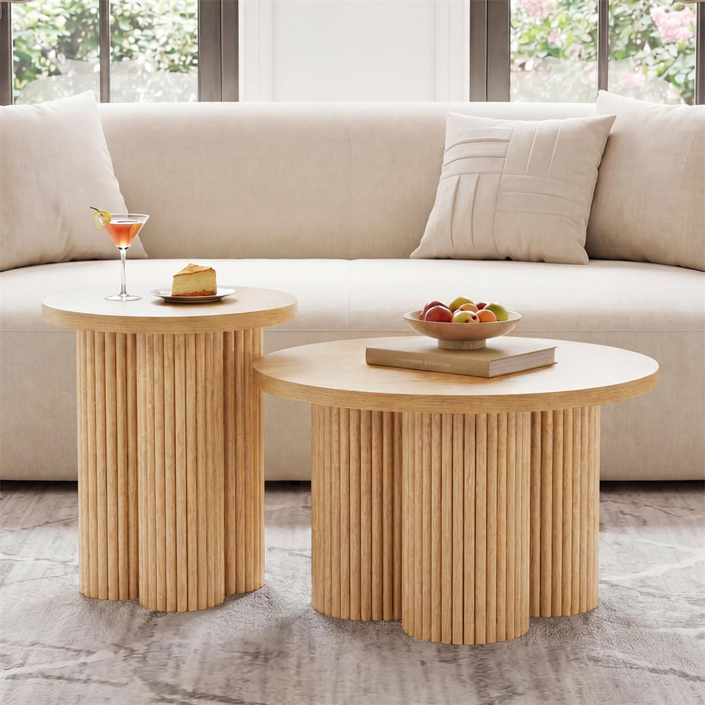 Two round nesting coffee tables with fluted pedestal bases in a warm light wood finish, styled in a bright living room with a beige sofa and decorative items on top.