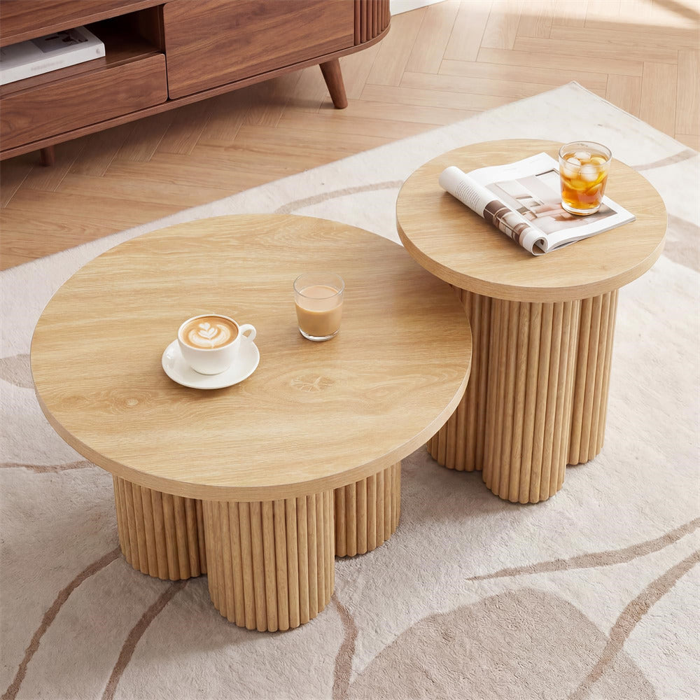 Two round nesting side tables with light natural wood tops and ribbed cylindrical pedestal bases, styled on a beige patterned rug with a coffee cup, drink, and magazine on top.