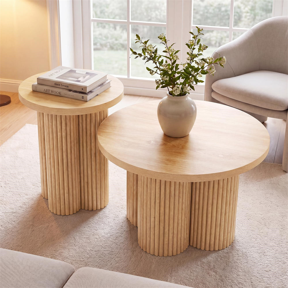 Two round nesting coffee tables with fluted pedestal bases in a bright, modern living room. The larger table is in the foreground and holds a beige ceramic vase with green branches, while the smaller table in the background holds a stack of three books.