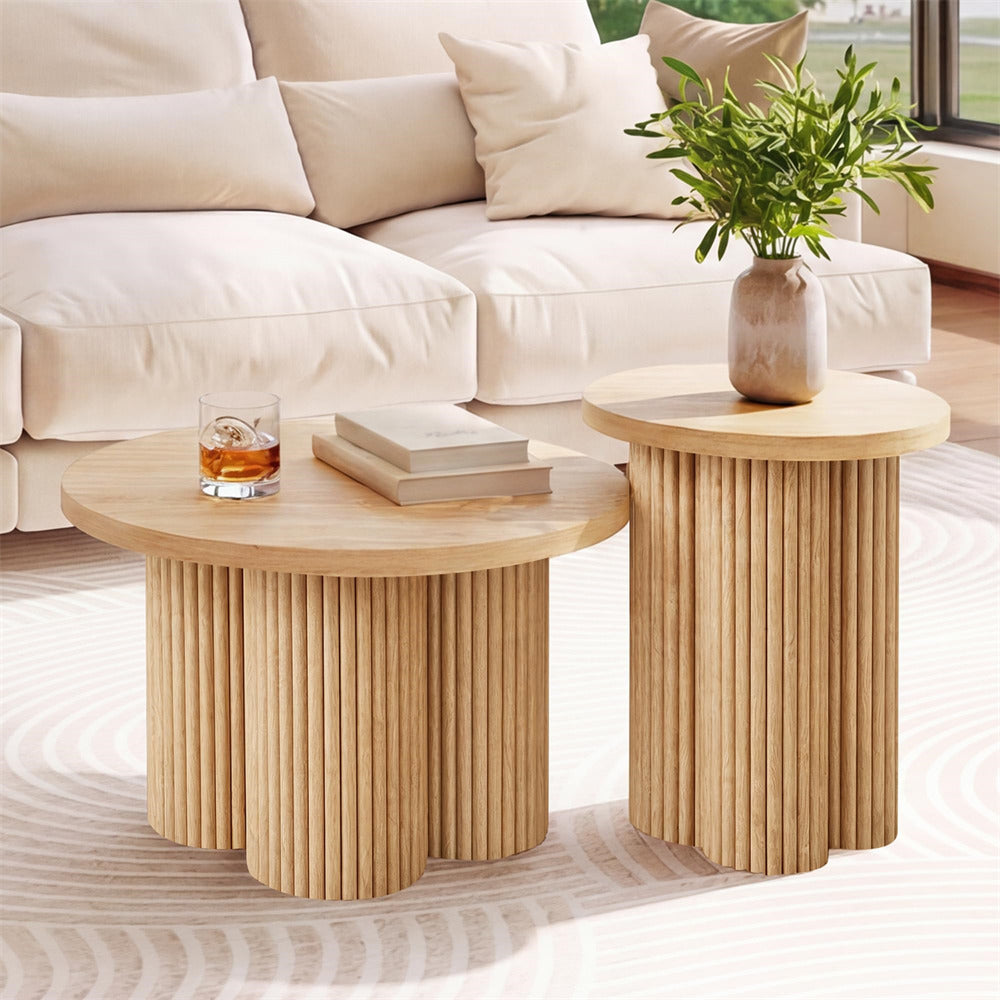 Two round wooden accent tables with fluted pedestal bases in a bright living room. One table holds a glass of whiskey and a stack of books, and the other holds a potted green plant.