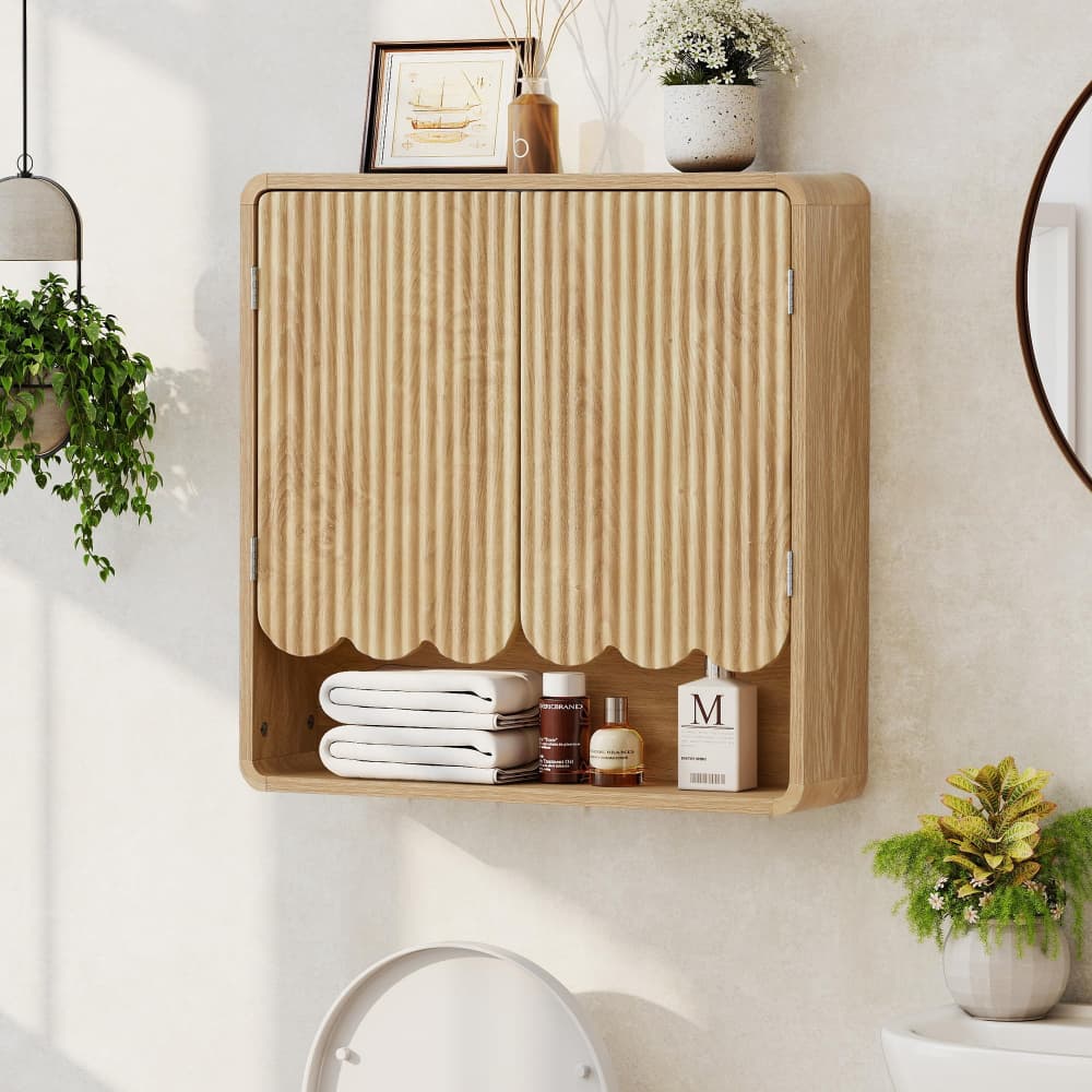 A light natural wood wall-mounted bathroom cabinet with two vertically slatted doors and an open lower shelf, styled with folded white towels, bottles, and small decor items against a warm off-white wall.