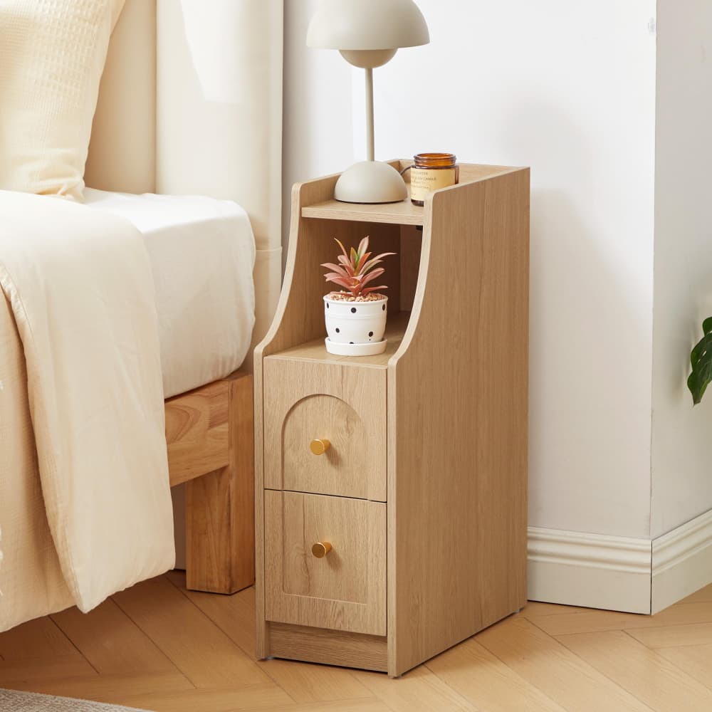 A light oak wood bedside table with two drawers and an open upper shelf, styled next to a bed in a bright bedroom. The table features gold-finished drawer knobs and holds a small potted succulent and a white table lamp.