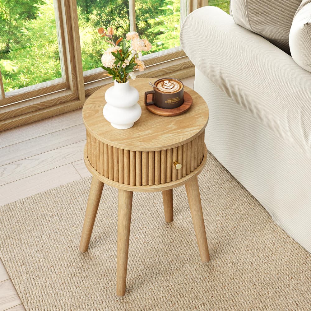 A round light oak wood side table with three tapered legs and a slatted cylindrical drawer front, styled in a bright living room next to a sofa with a coffee cup and flowers on top.