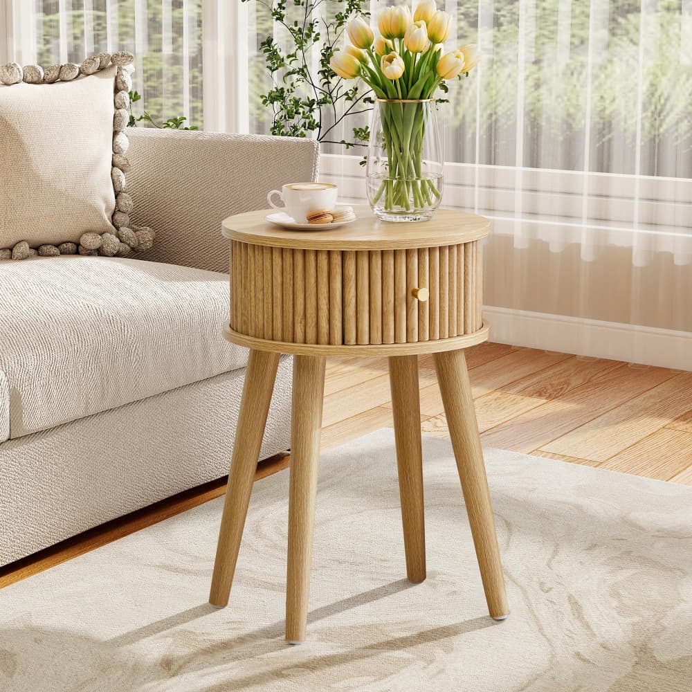 A light natural wood side table with a single slatted drawer front and three tapered legs, styled in a bright living room next to a sofa with a cup and flowers on top.