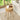 A round light oak wood side table with three tapered legs and a slatted cylindrical drawer front, styled in a bright living room next to a sofa with a coffee cup and flowers on top.