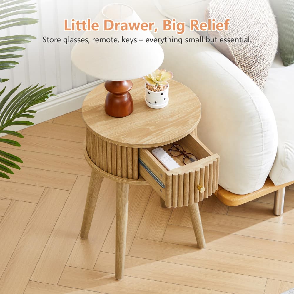 A light wood round side table with a slatted front and legs, featuring an open drawer holding glasses and a remote. A white table lamp and a small decorative pot sit on top.