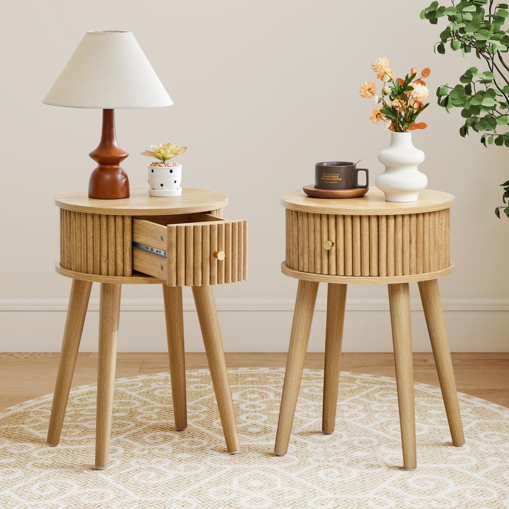 Two light natural wood side tables with ribbed drawer fronts and tapered legs, styled in a room setting. One table holds a brown table lamp and a small white pot, while the other holds a white vase with flowers and a dark brown cup.