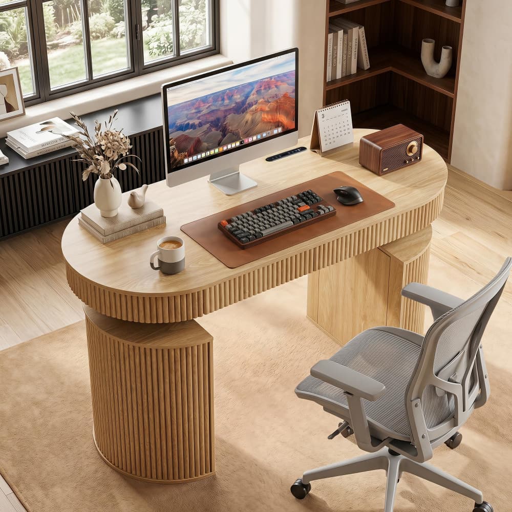 A modern curved wooden home office desk with ribbed cylindrical legs, styled with a computer monitor, keyboard, mouse, books, and a coffee mug, next to a gray mesh office chair in a bright room.