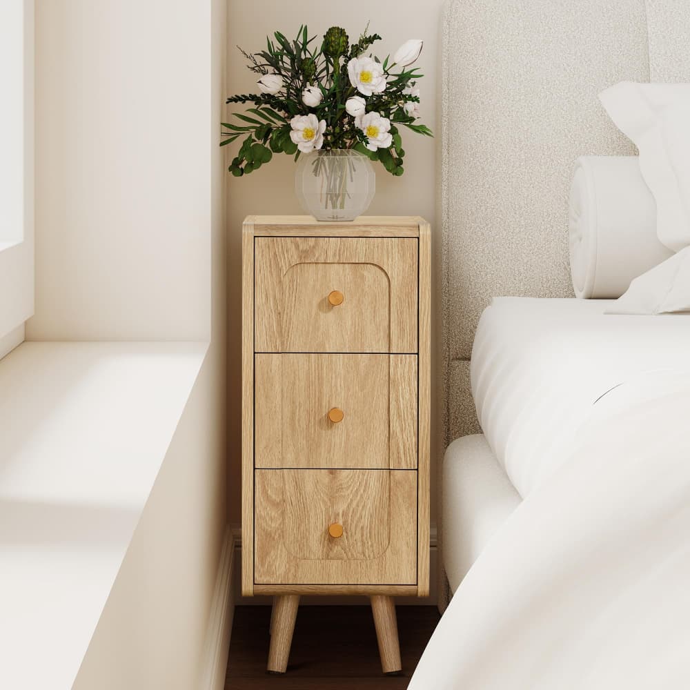 Wooden nightstand with three drawers next to a bed with white bedding.