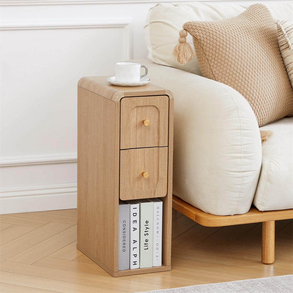 A light oak wood two-drawer nightstand with small round knobs, styled next to a cream sofa with a cushion and books on a light wood floor.
