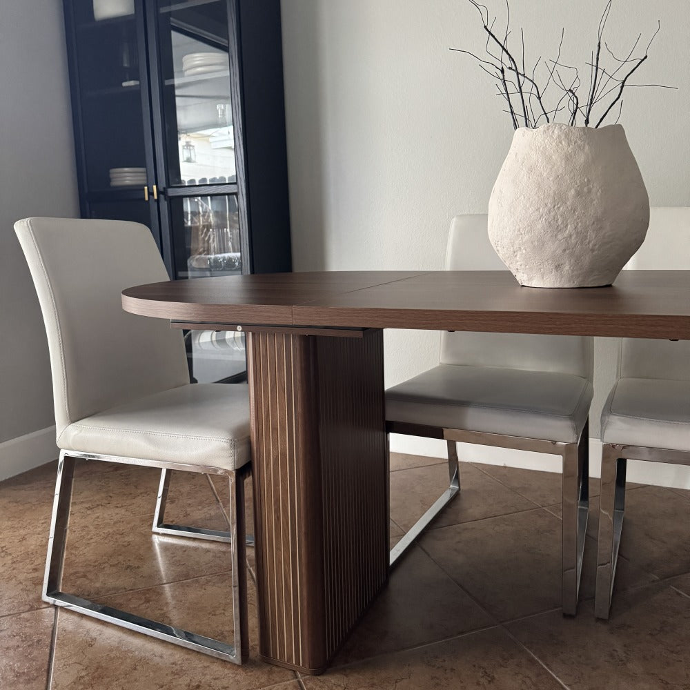 Dining room with a wooden table and white chairs, featuring a vase with branches on the table.