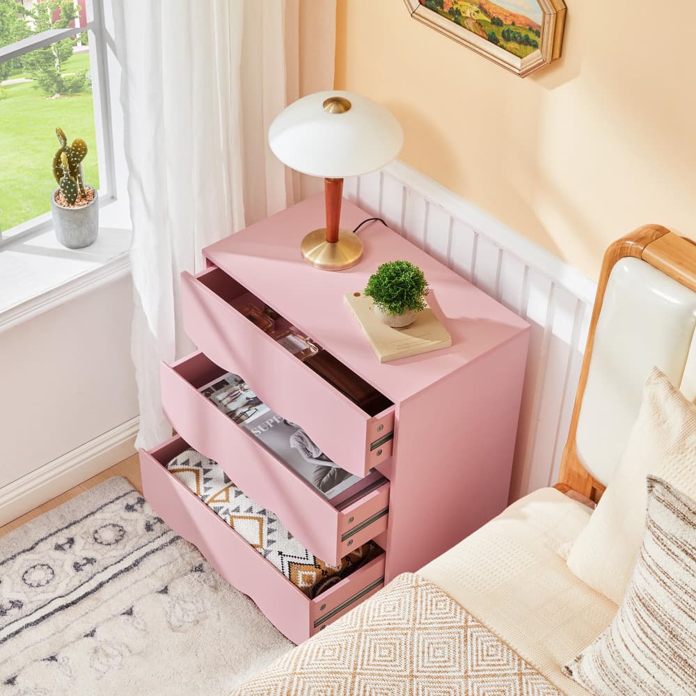 A pink three-drawer nightstand in a cozy bedroom setting, with two drawers open to show storage space. The nightstand is styled with a white table lamp and a small potted plant, next to a bed with neutral-toned bedding.