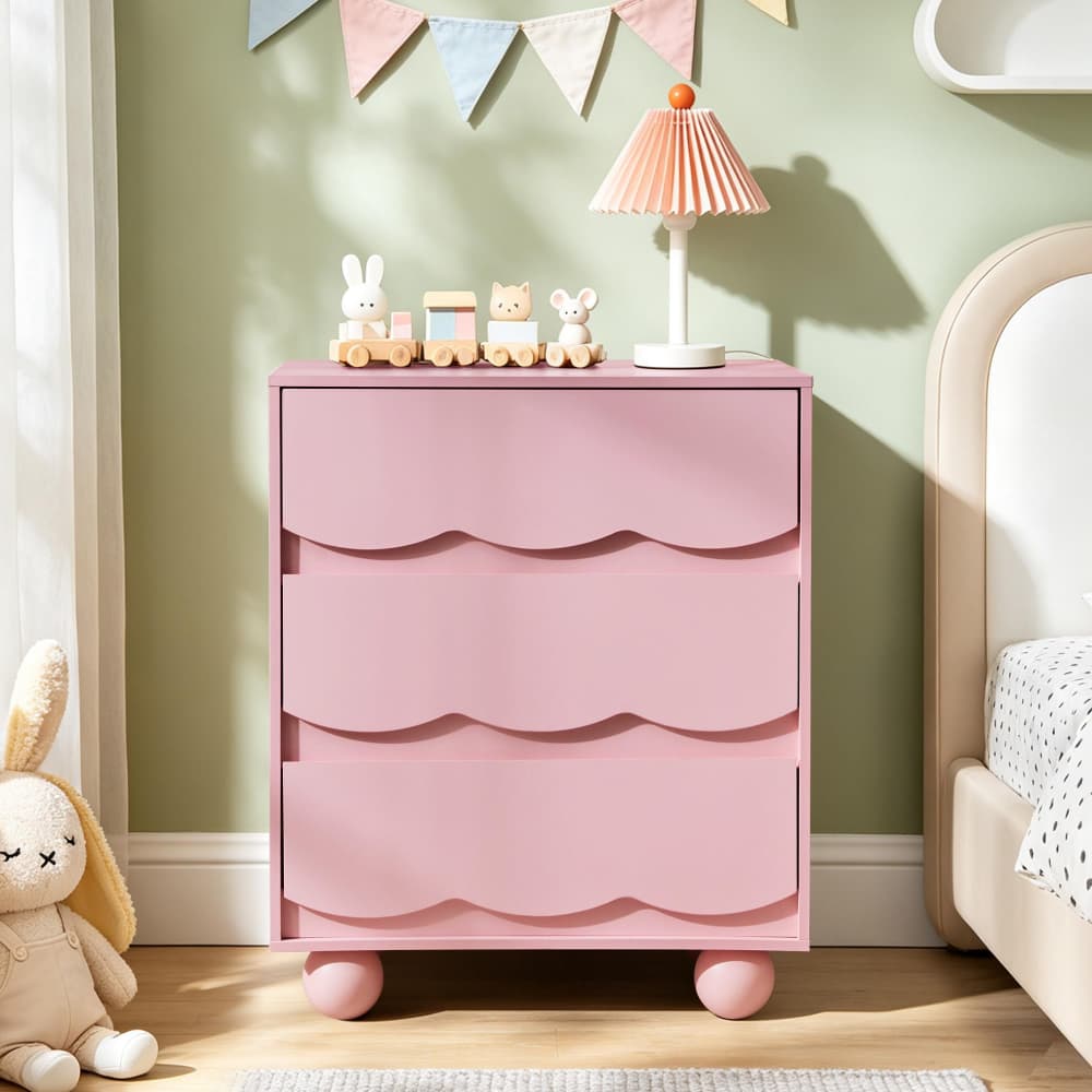 A pink three-drawer nightstand with scalloped drawer fronts and rounded feet, styled in a child's bedroom next to a bed with a pleated pink lamp and small toy train decorations on top.