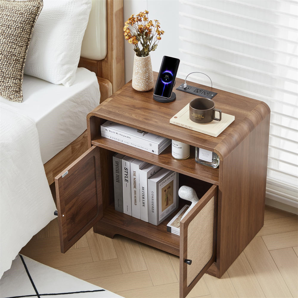 Wooden nightstand with open drawer, books, a mug, and a phone on top next to a bed.
