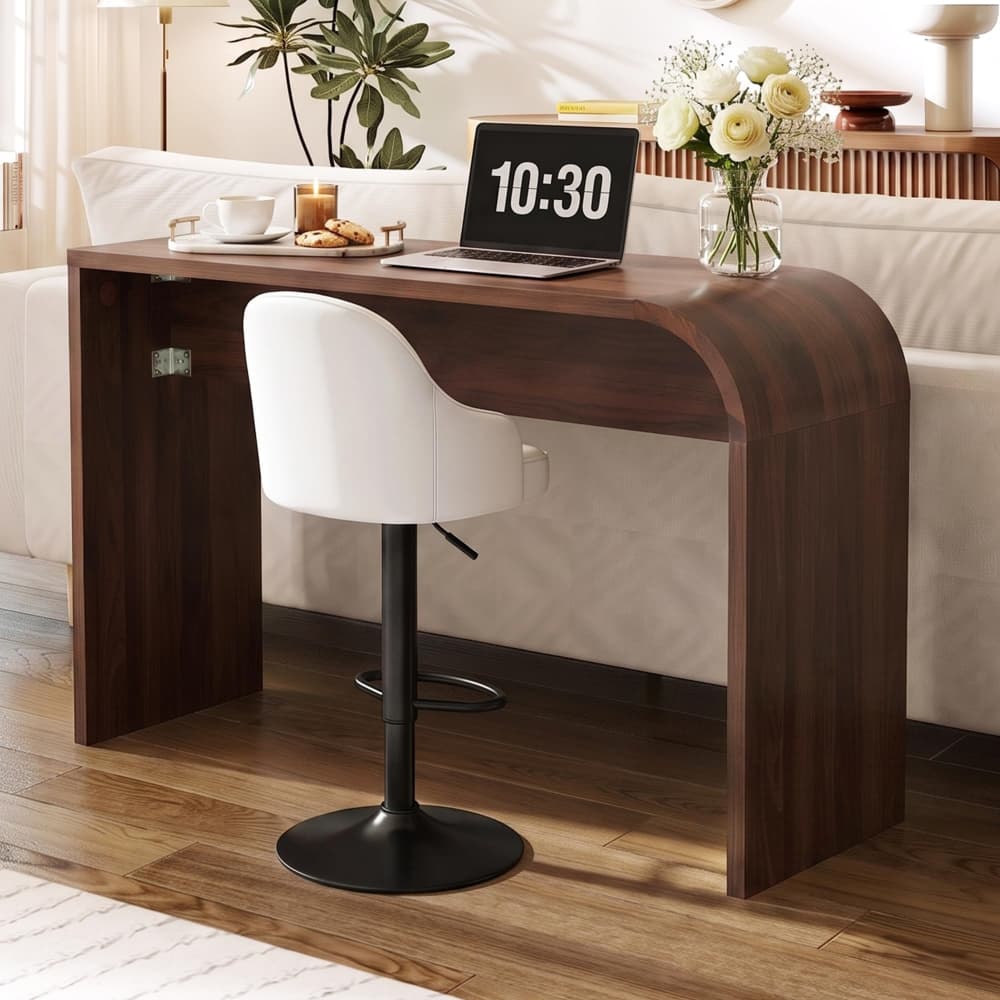 A modern curved wooden console table with a dark brown finish, paired with a white swivel barstool with a black pedestal base. The table is styled in a bright living room setting with a laptop, flowers, and a coffee cup on top.