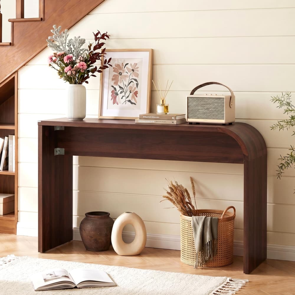 A dark brown wooden console table styled in a home entryway setting, holding a white vase with pink and burgundy flowers, a framed botanical print, books, a retro radio, and decorative vases on a light wood floor against a cream shiplap wall.