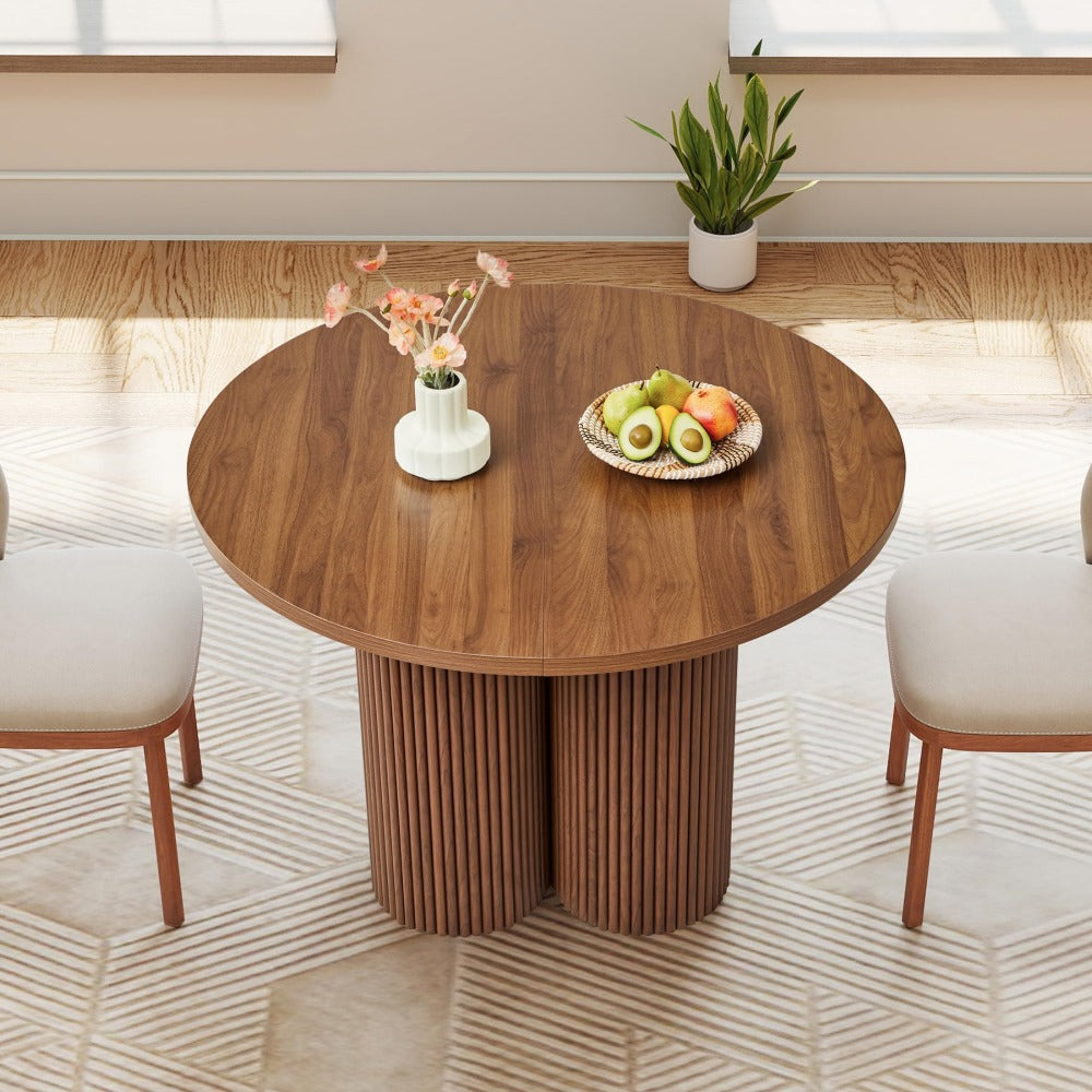 A round wooden dining table with a fluted pedestal base, styled in a bright dining room with two cream chairs, a fruit bowl, and a small vase.