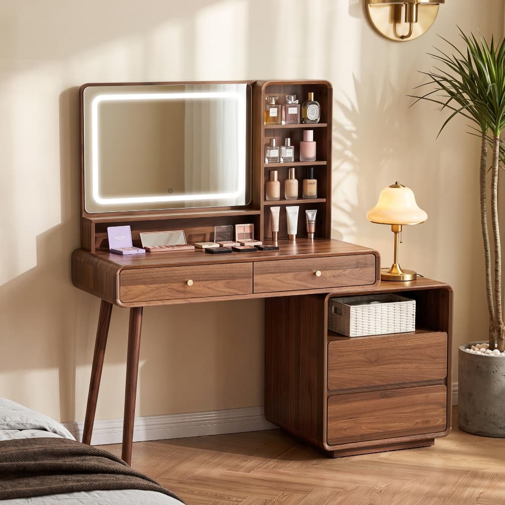 A walnut wood vanity table with an illuminated rectangular mirror, open shelving, two drawers, and a matching side cabinet, styled in a warm beige bedroom setting with a lamp, plant, and bed visible.