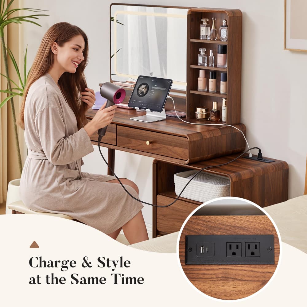 A woman sits at a wooden vanity desk with a mirror, charging accessories, and a hair dryer in a bright room. The desk has built-in storage shelves and a circular inset showing a power outlet panel on the wood surface.