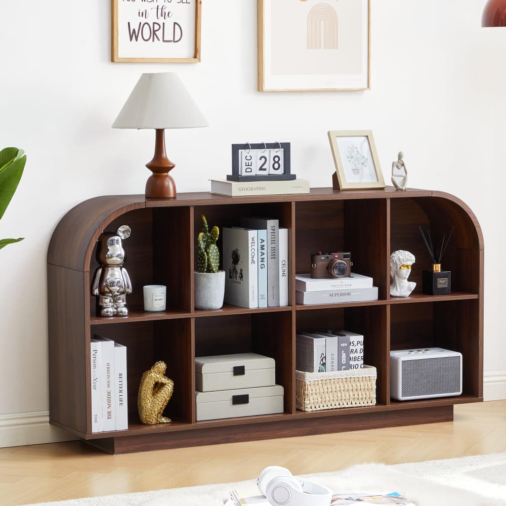 A dark brown wooden arched bookcase shelf styled with books, a lamp, small figurines, a cactus, and storage boxes in a bright, modern room.