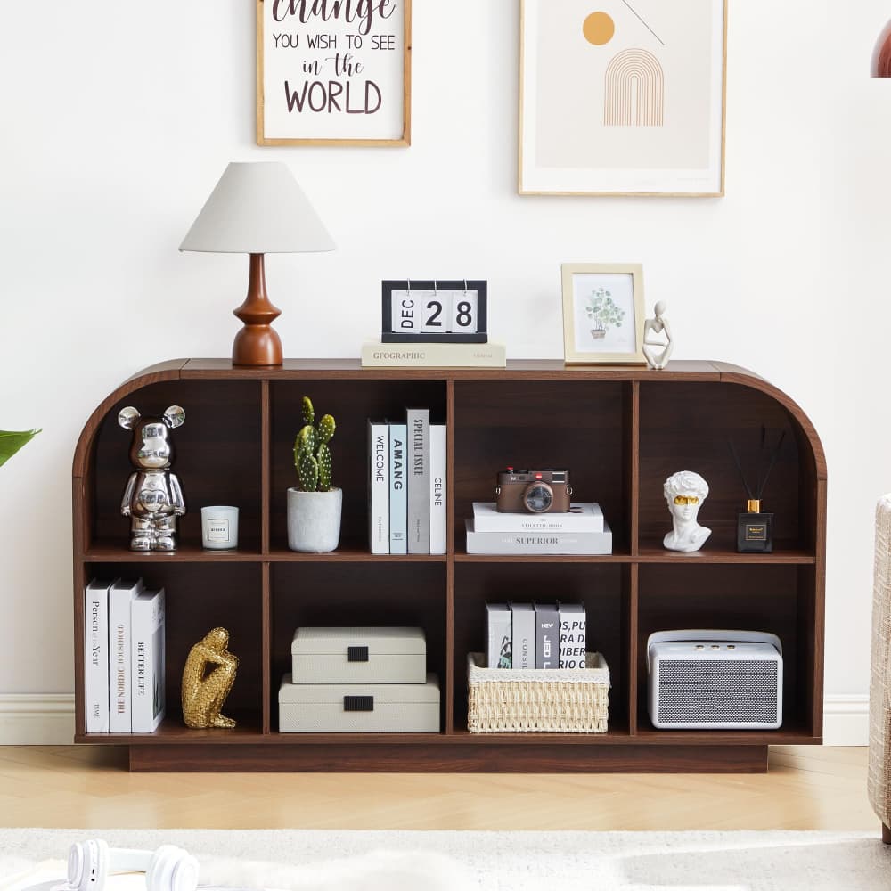 A dark brown wooden console shelf with a curved top and multiple cubbies, styled with books, a lamp, a cactus, a camera, and decorative objects in a bright room.