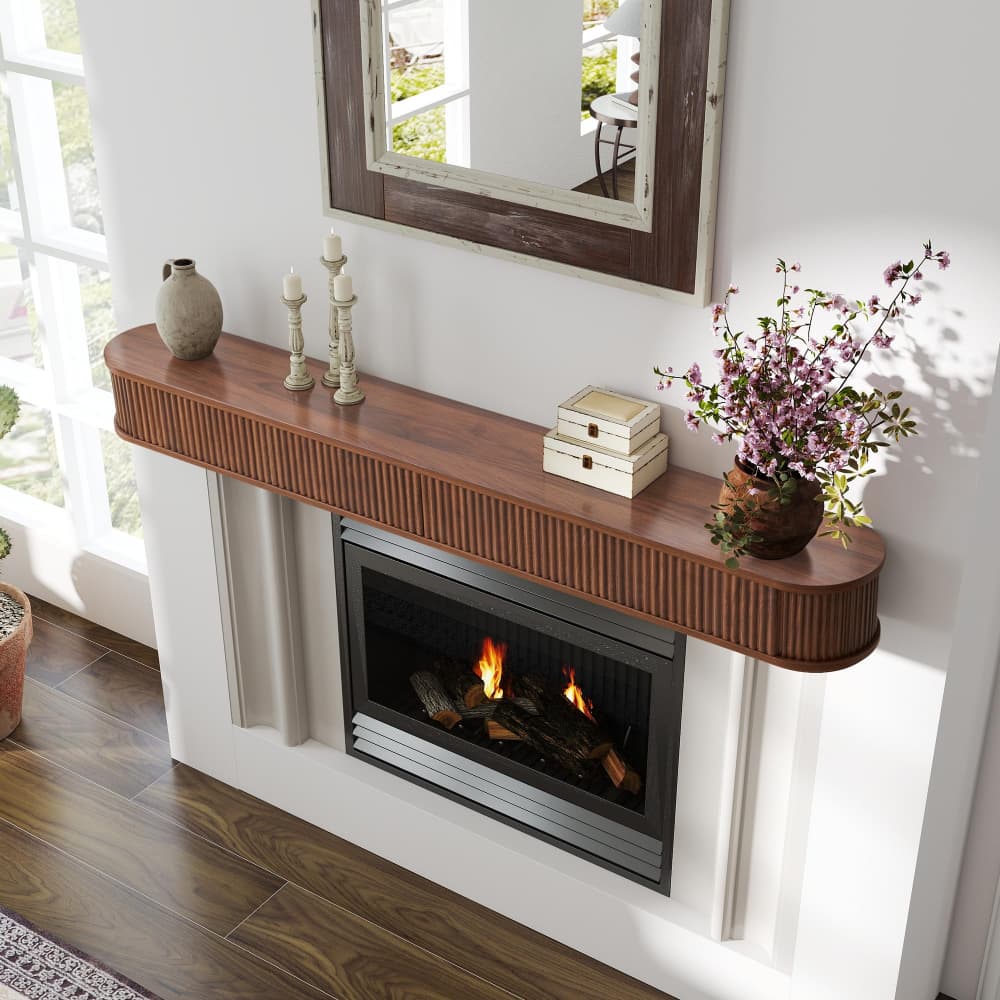 A curved wooden mantel shelf mounted above a modern electric fireplace in a white fireplace surround, styled with candles, a vase, stacked books, and a potted plant in a bright room.