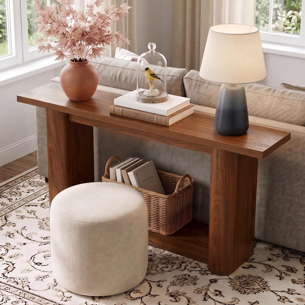 A wooden console table in a bright living room setting, styled with a terracotta vase of dried pink flowers, a glass cloche, a small bird figurine on stacked books, a gray-blue gradient table lamp, and a round off-white ottoman in front.