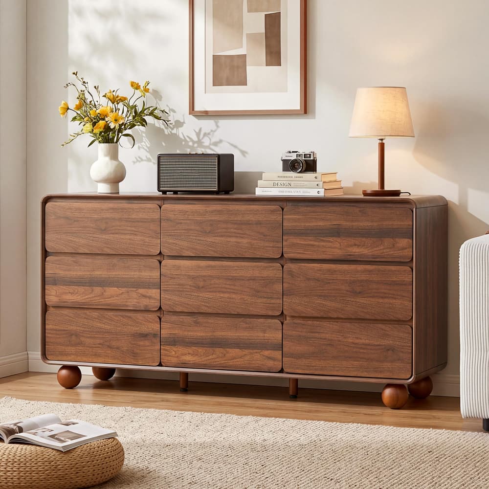 A mid-century modern wooden dresser with nine drawers in a warm walnut finish, styled in a cozy living room setting with a lamp, radio, and flowers on top.