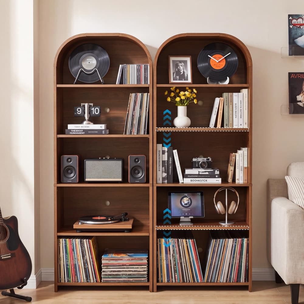 Two tall wooden bookshelves with arched top sections, styled with vinyl records, books, speakers, a record player, headphones, and decor, set in a bright living room next to a sofa.