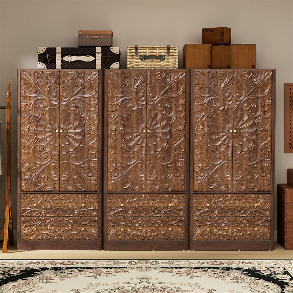 A large carved wooden wardrobe or cabinet with three doors and three drawers, featuring intricate floral relief carvings and brass-tone knobs, shown in a styled room setting with vintage trunks and a patterned rug.