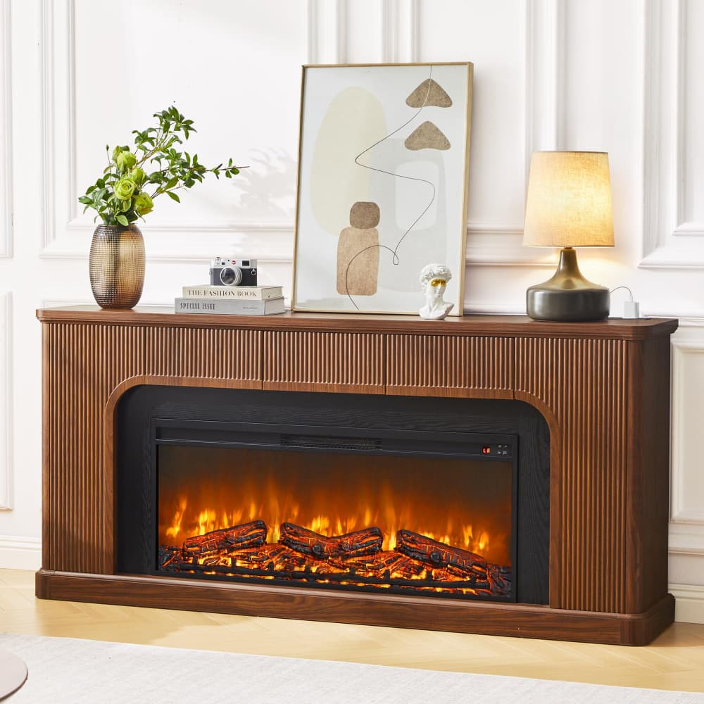 A brown wood electric fireplace with a faux log fire burning inside, styled on a mantel with a vase of greenery, books, a framed print, and a table lamp against a white paneled wall.
