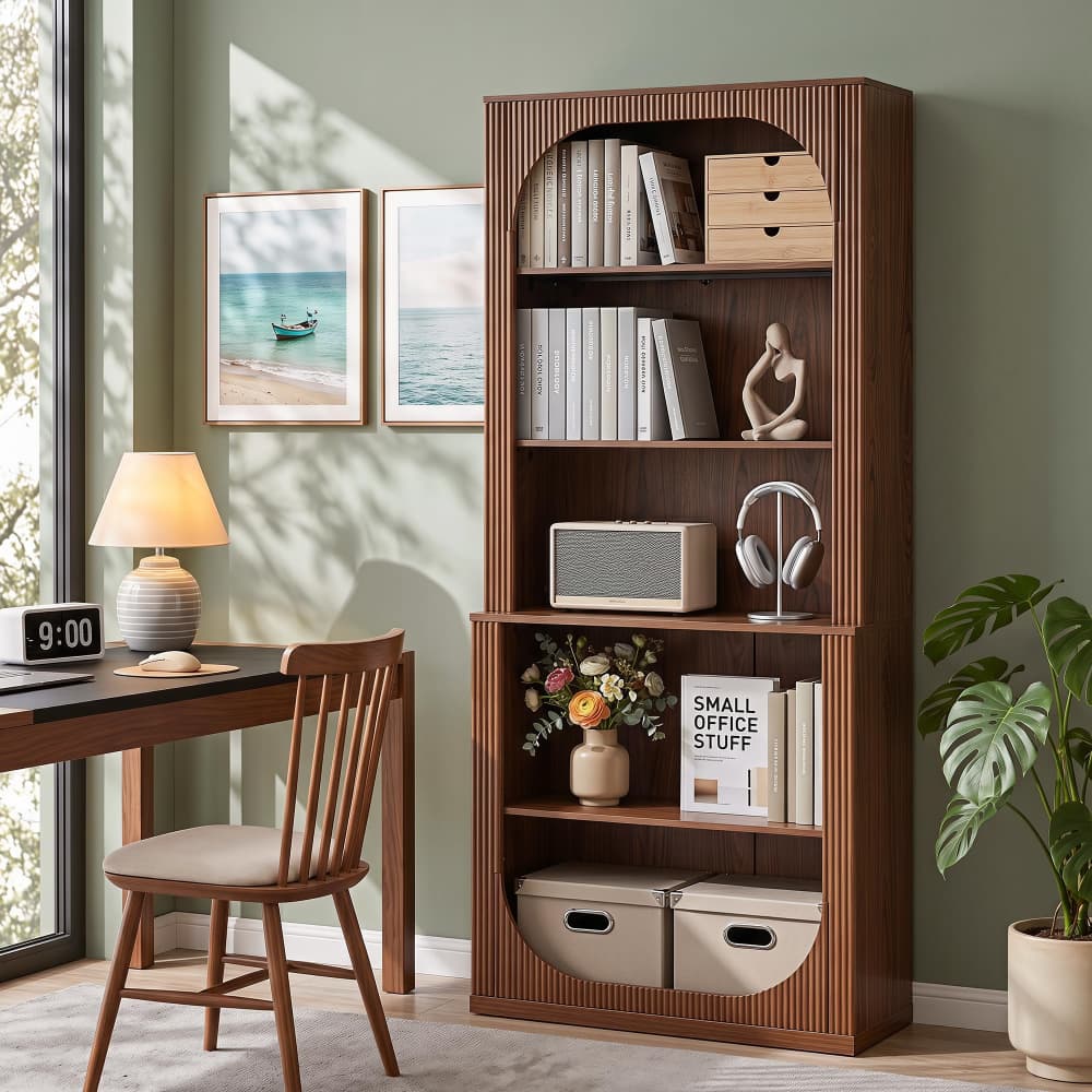 A tall wooden bookcase with a warm medium-brown finish stands in a styled home office. The bookcase features an arched top shelf, four open middle shelves, and two lower storage bins.
