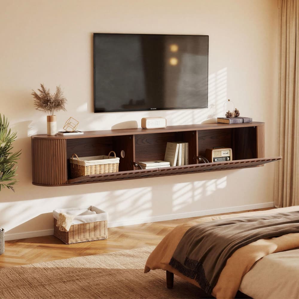 A floating dark wood wall-mounted TV stand and shelf unit in a styled bedroom, with a television mounted above it, books, baskets, and decor on the shelves, set against a warm beige wall with sunlight and shadows.