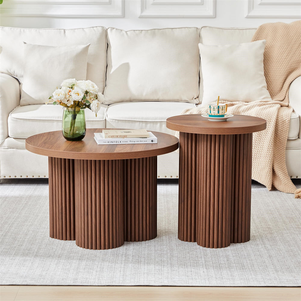 Two round wooden coffee tables with fluted pedestal bases in a bright living room. One table holds a green glass vase with white flowers and a book, and the other holds a small cup and saucer.