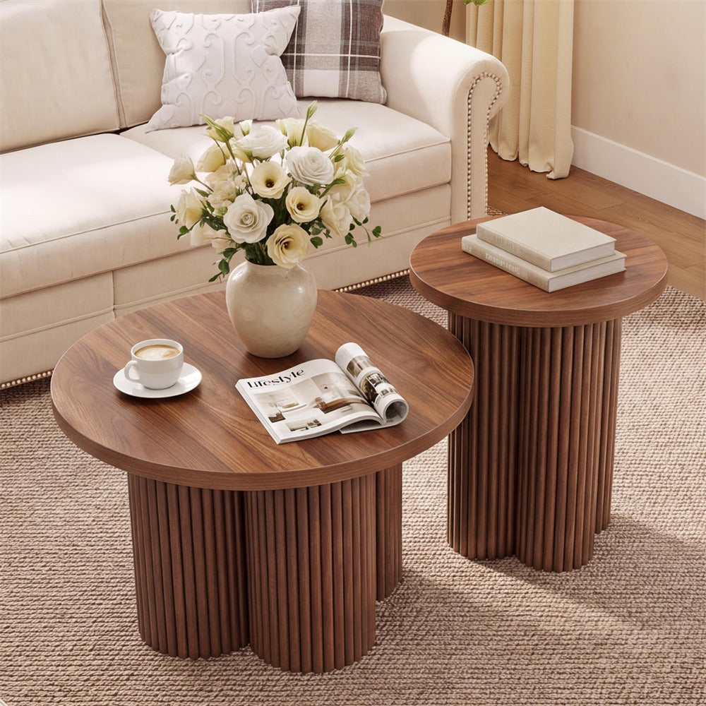 Two round wooden coffee tables with fluted pedestal bases in a styled living room. The larger table holds a vase of white flowers, a cup and saucer, and an open magazine, while the smaller matching table beside it holds two stacked books.