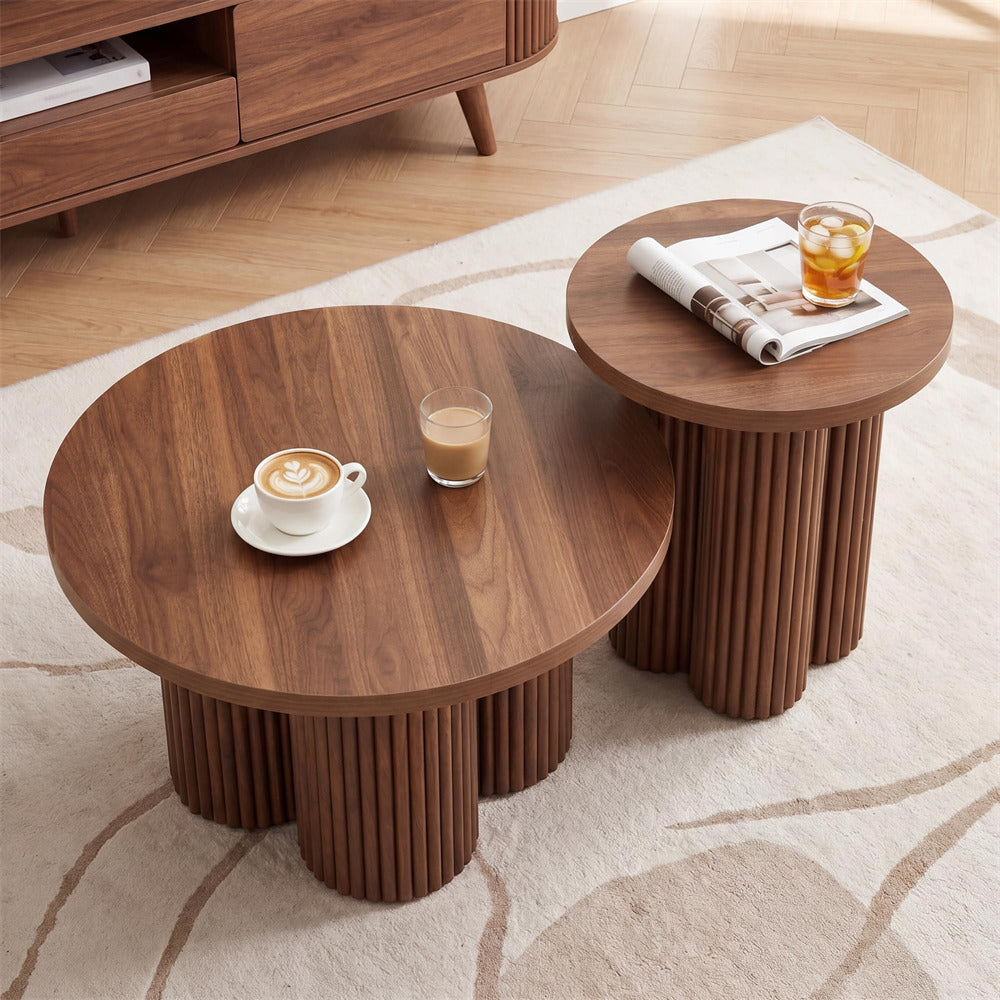 Two round nesting coffee tables with fluted pedestal bases in a warm medium brown wood finish, styled on a light patterned rug with a coffee cup, a glass of milk coffee, and a rolled magazine on the smaller table.
