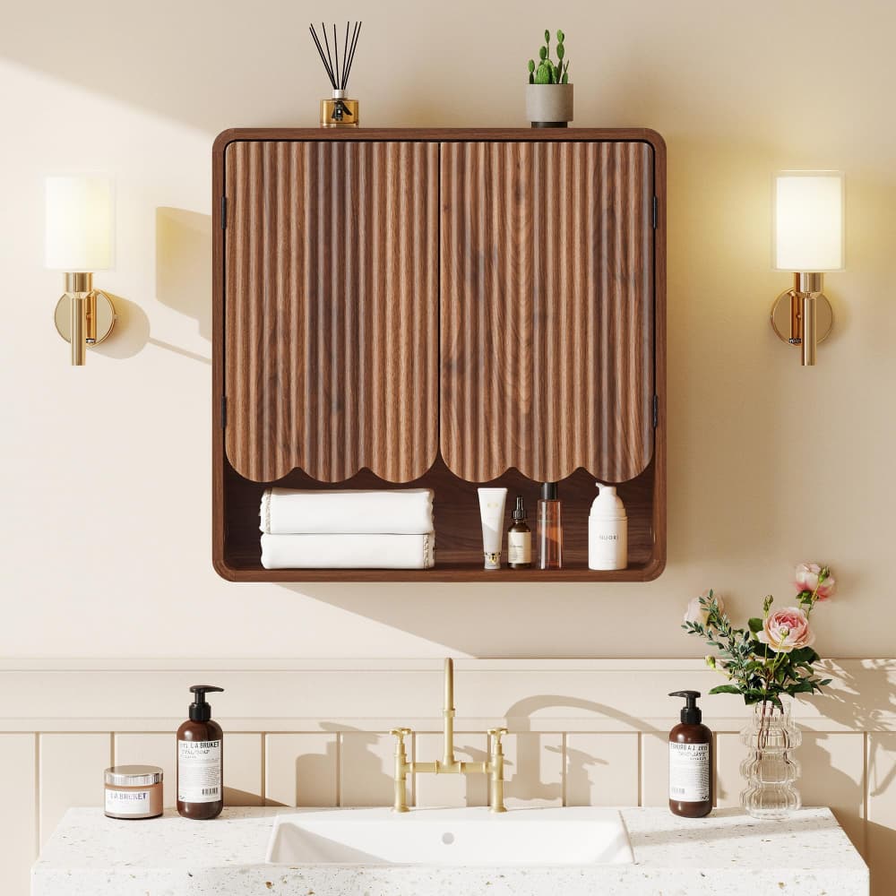 A wall-mounted bathroom cabinet with two vertically slatted wood doors in a warm medium brown finish, shown installed above a sink in a styled bathroom. The cabinet has a lower open shelf holding folded white towels and small toiletries, and is flanked by wall sconces and decorative items.