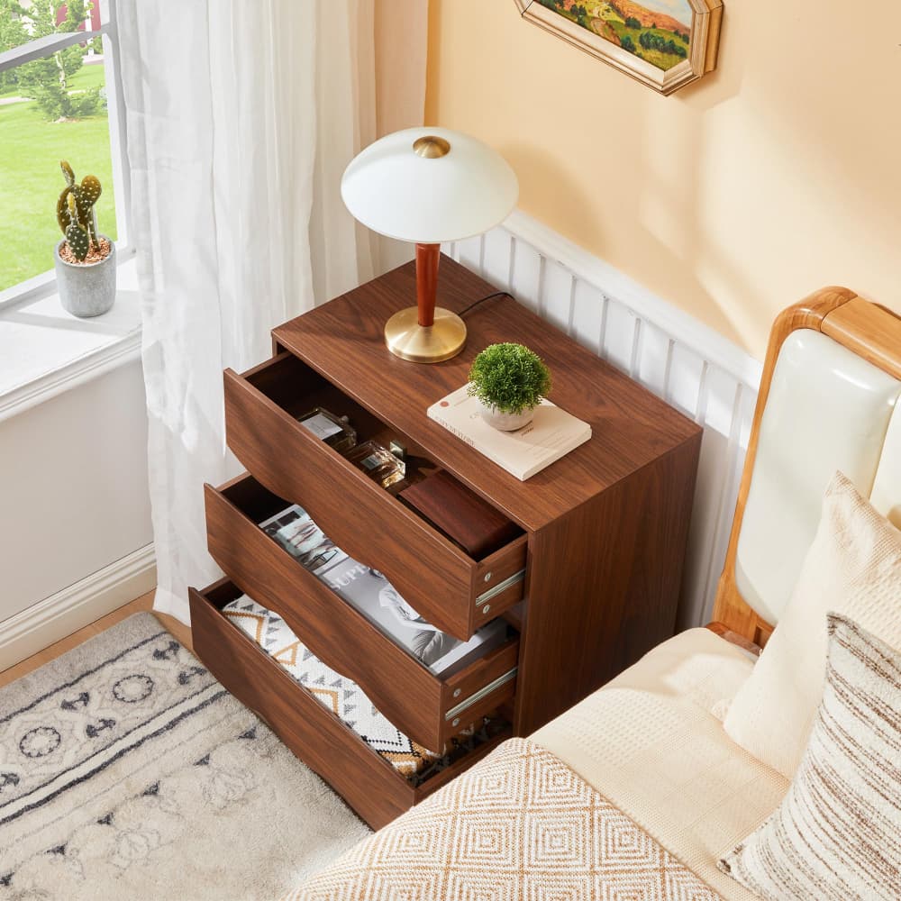 A three-drawer wooden nightstand with a warm walnut finish, styled in a bedroom next to a bed. The drawers are open, revealing magazines and folded linens inside, while a white table lamp and a small potted green plant sit on top.