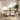 A mid-century modern dining set featuring a round wooden table and six matching chairs with white upholstered seats and wooden frames, styled in a bright, contemporary dining room.