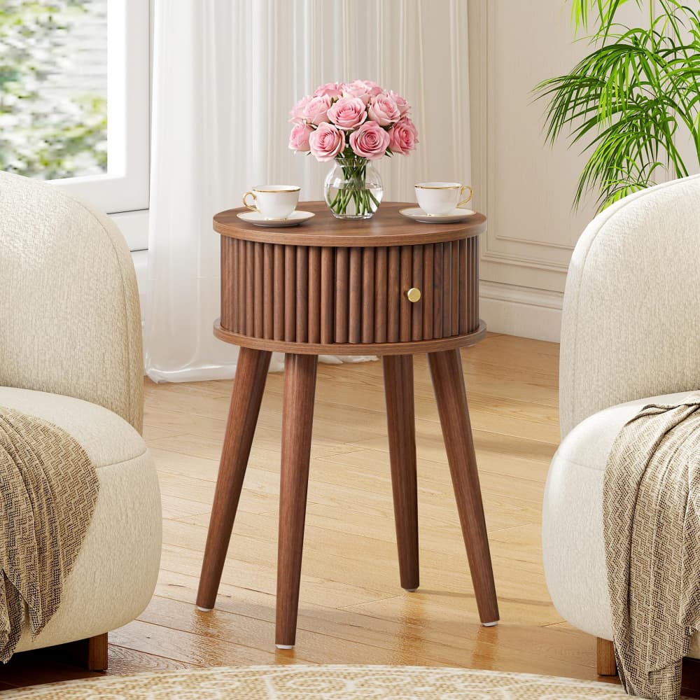 A round wooden side table with a slatted front and four tapered legs, styled in a bright living room between two cream armchairs. The table holds a vase of pink roses and two white teacups with saucers.
