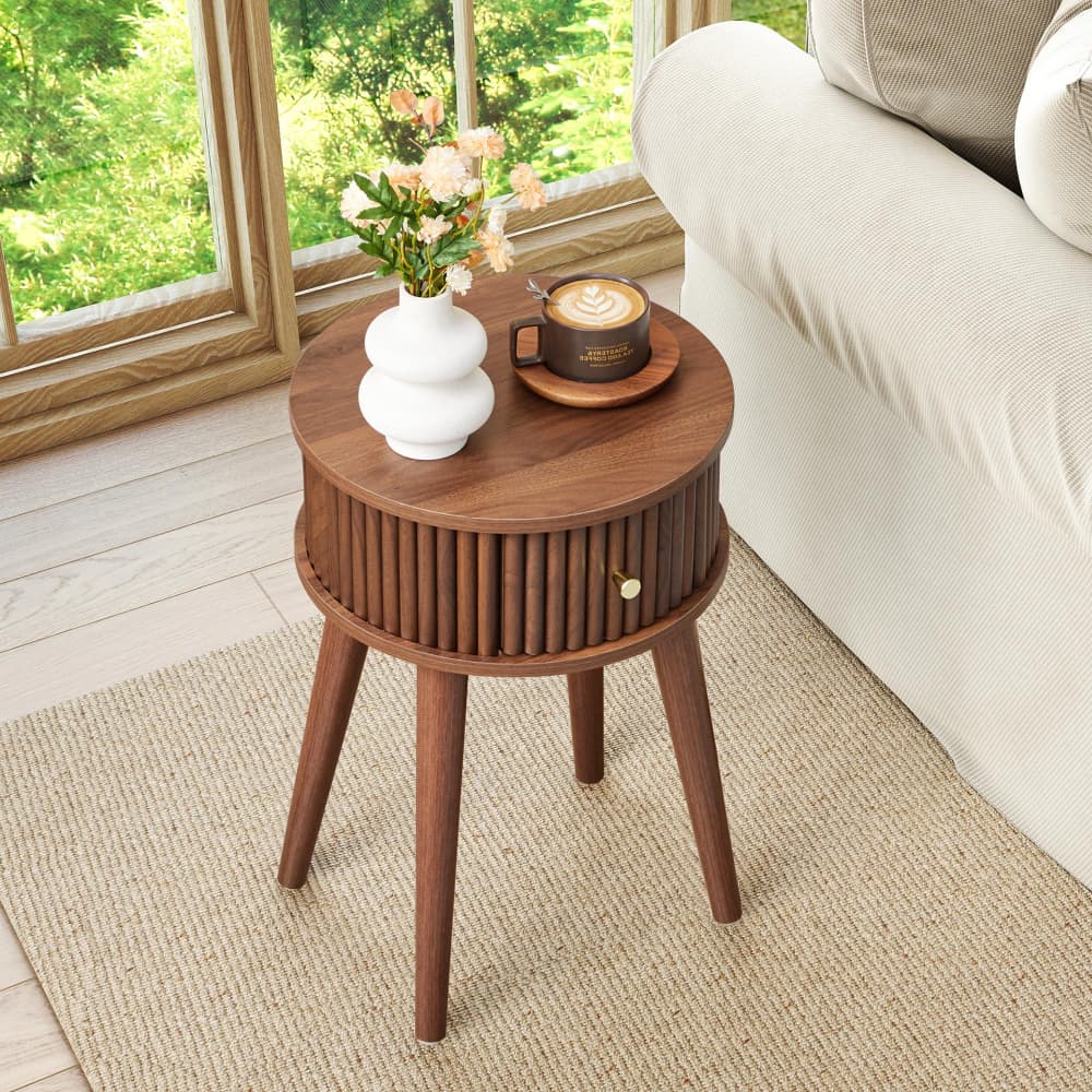 A round wooden side table with three legs and a slatted drawer front, styled in a bright living room next to a sofa with a vase of flowers and a cup of coffee on top.