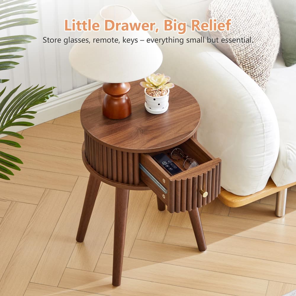 A round wooden side table with a slatted front and three legs, shown beside a white sofa in a bright room. The table has one open drawer and is styled with a white lamp and a small potted succulent.