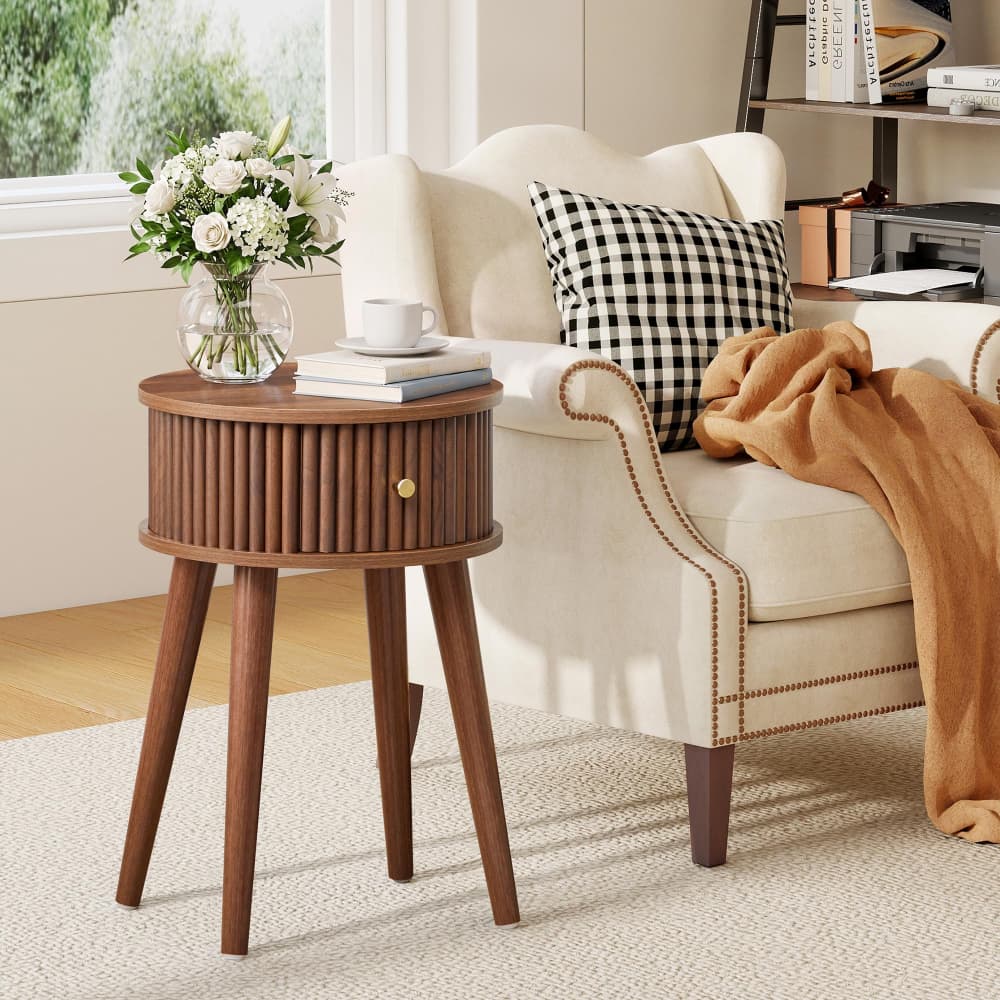 A round wooden end table with three tapered legs and a ribbed front drawer, styled in a bright living room next to a cream armchair with a black-and-white check pillow and a tan throw blanket.