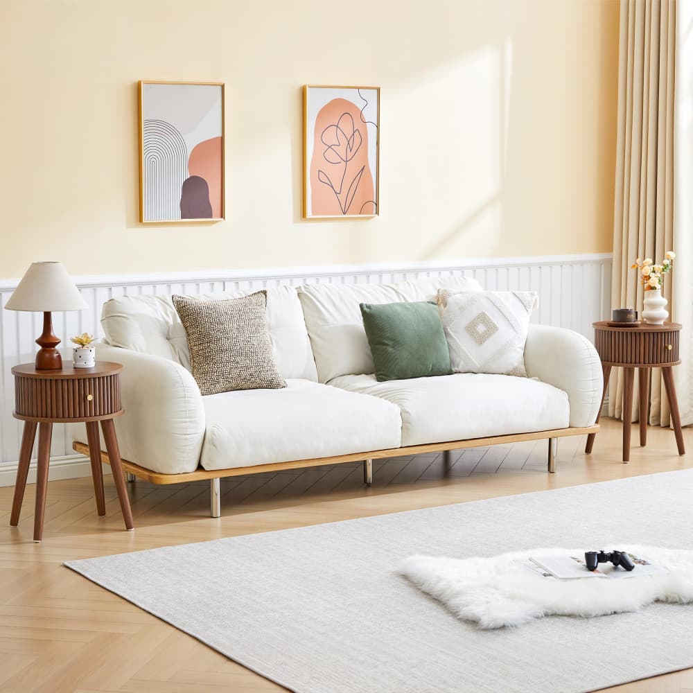 A cream-colored fabric sofa with a light wood frame and legs, styled in a warm, modern living room with beige walls, decorative pillows, side tables, and a light rug.