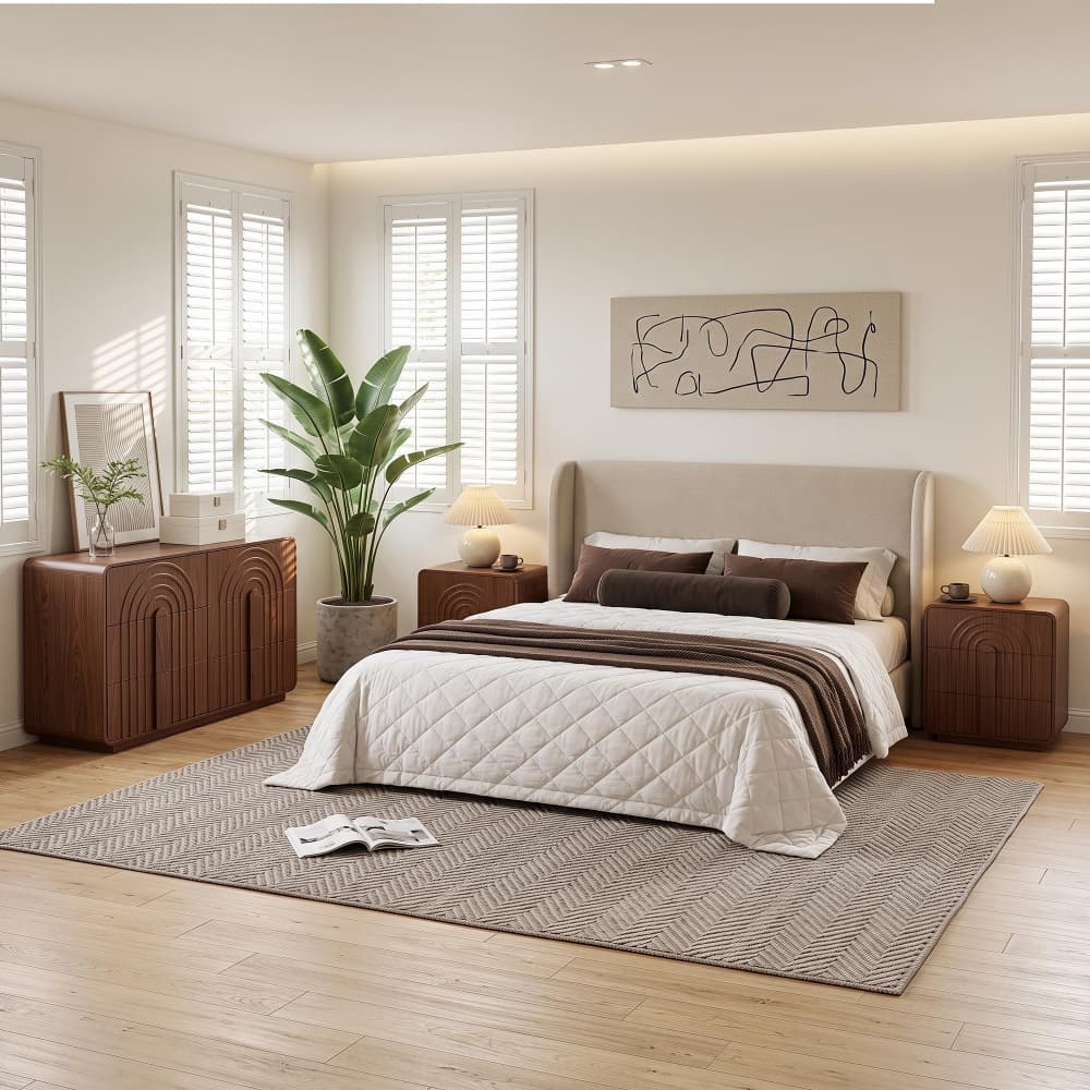 A styled bedroom interior featuring a bed dressed in white and brown bedding, flanked by matching wood nightstands, with a wood dresser, a large potted plant, and a gray patterned rug on a light wood floor.