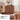 A mid-century modern wooden side cabinet with a warm brown finish, featuring a sculpted geometric arch design on the front. The cabinet is styled in a bright living room next to a beige armchair, with a vase of flowers and a book on top.