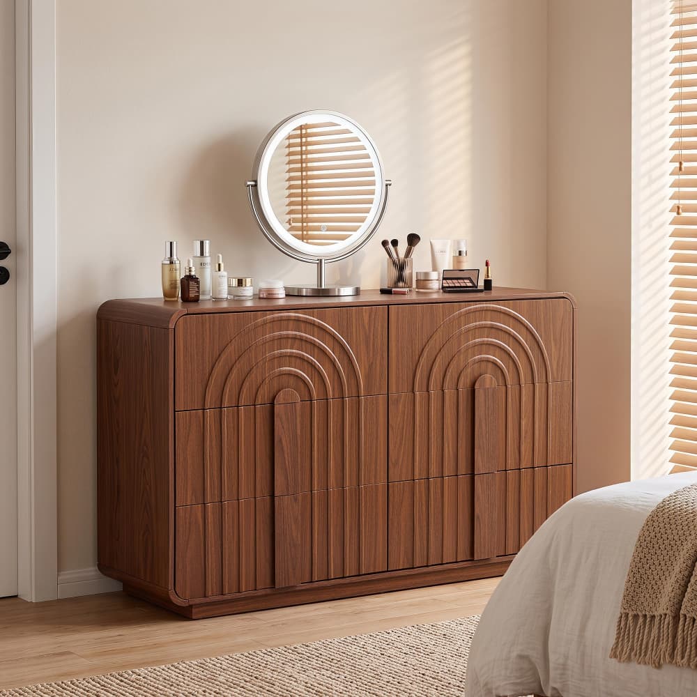 A wooden vanity cabinet with arched panel detailing and a round illuminated mirror on top, styled in a bedroom setting next to a bed.