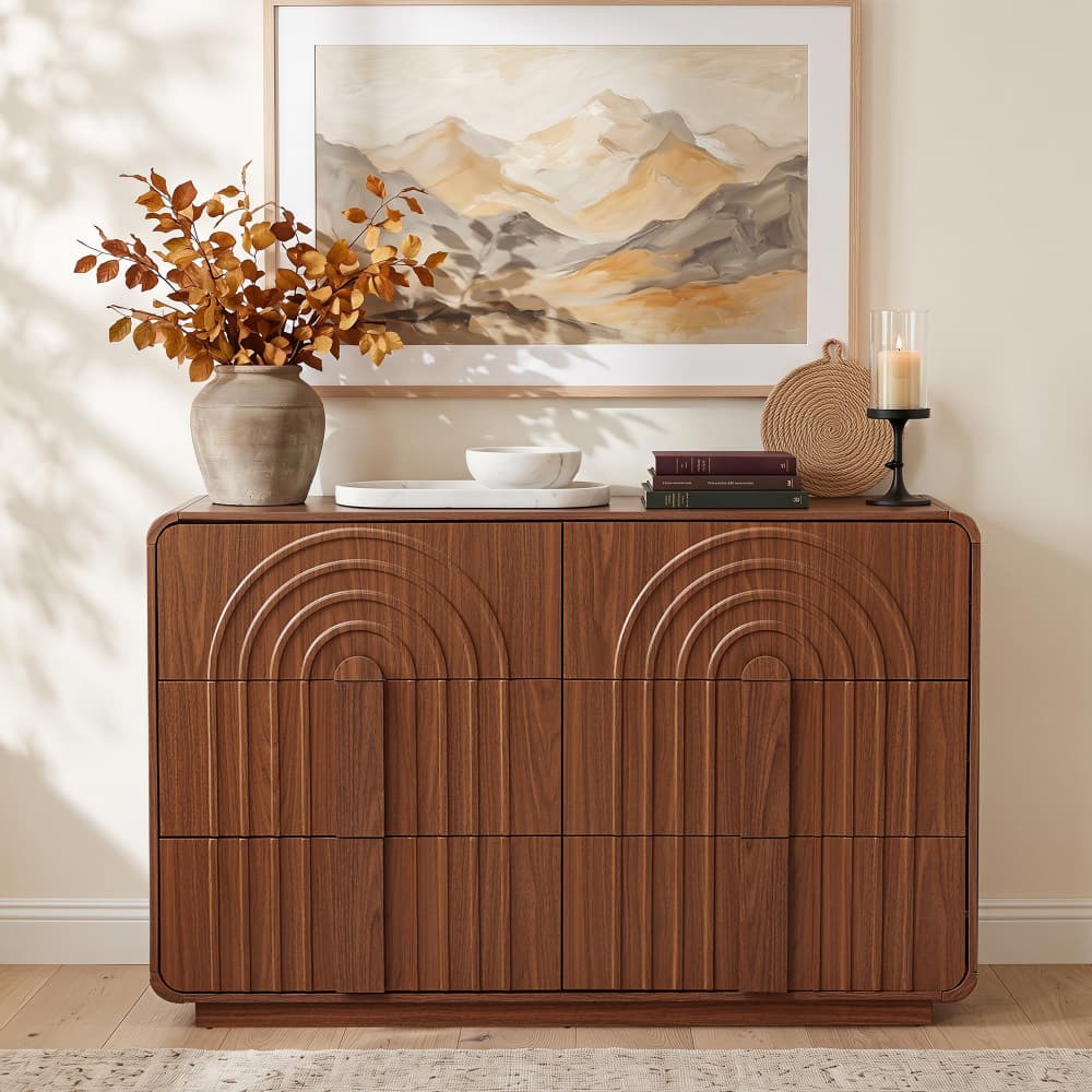 A mid-century modern wooden credenza or sideboard with a warm walnut finish, featuring a curved arched panel design on the front. The cabinet is styled in a living room setting with a vase of dried leaves, a bowl, books, and a candle, beneath a framed mountain landscape print.